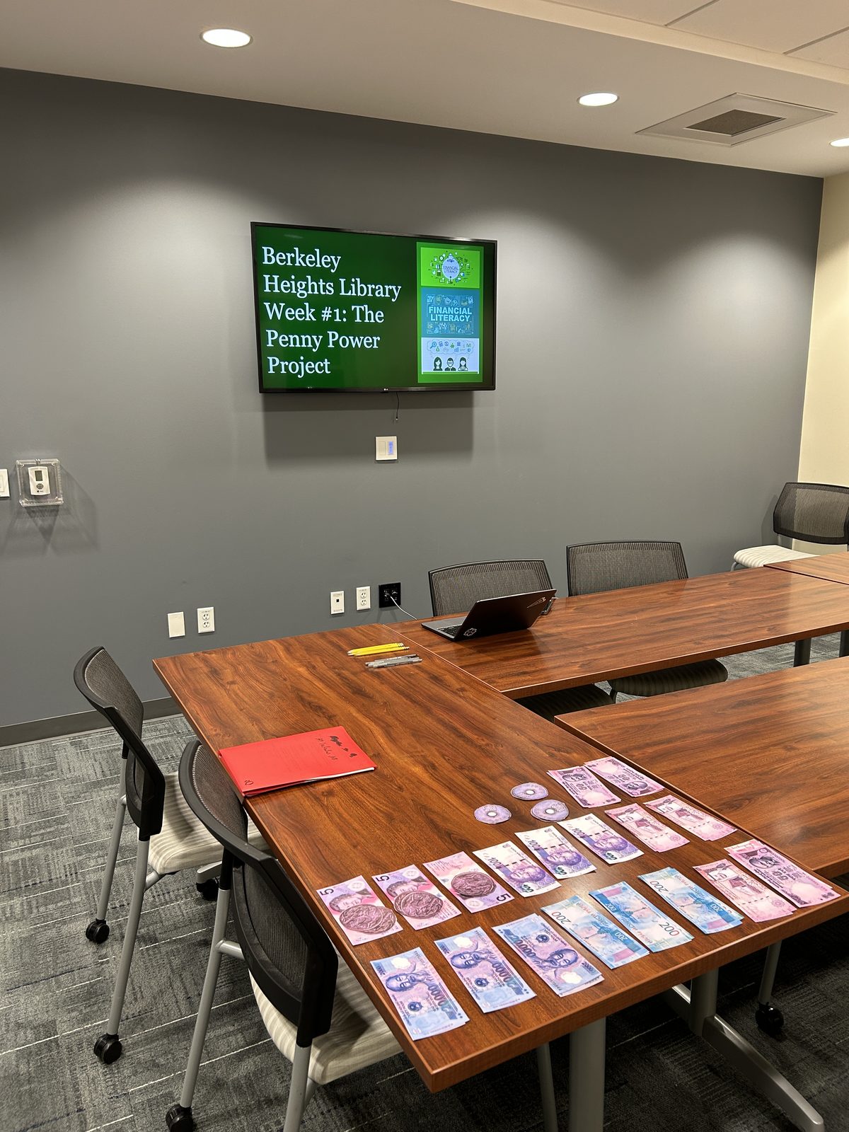 Penny Power Project slide on the TV at Berkeley Heights Library, with world currencies laid out on the table
